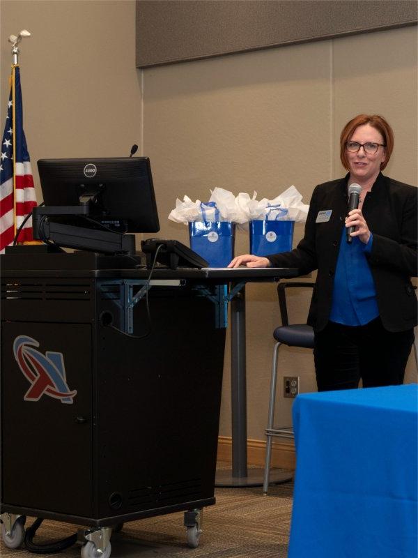 Kara Van Dam with microphone at front of room, hand resting on computer stand with blue gift bags behind her